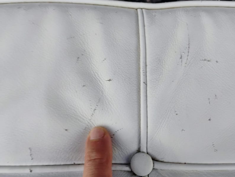 May include: Close-up of a white leather surface with visible wear and tear, including small dark spots and scratches. The surface is divided into sections by stitched seams and a button is visible.