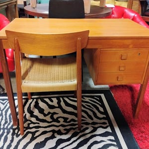 May include: A light brown wooden desk with three drawers and a matching chair. The chair has a woven seat. The desk and chair are set on a black and white zebra-print rug and a red rug.