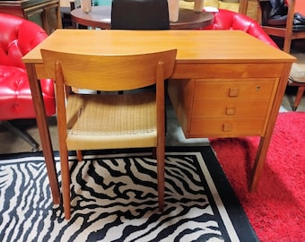 Könnte beinhalten: Ein hellbrauner Holzschreibtisch mit drei Schubladen und einem passenden Stuhl. Der Stuhl hat einen gewebten Sitz. Der Schreibtisch und der Stuhl stehen auf einem schwarz-weiß gestreiften Teppich und einem roten Teppich.