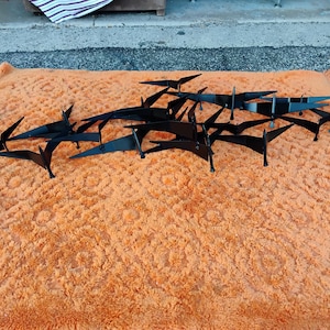 May include: Black metal wall art featuring a flock of stylized birds in flight. The birds are arranged in a dynamic composition, resting on an orange, textured surface. The artwork is designed to be mounted on a wall.