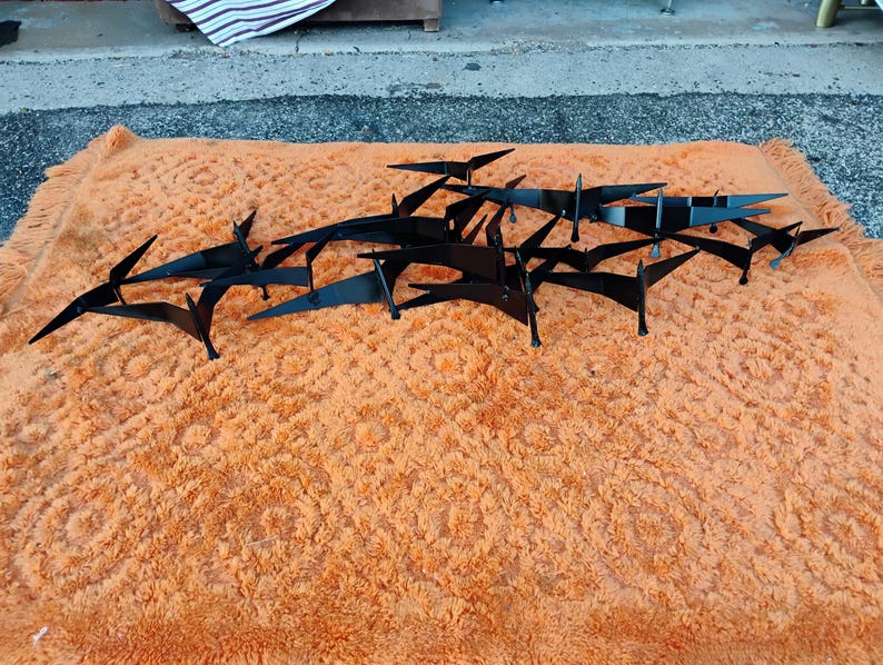 May include: Black metal wall art featuring a flock of stylized birds in flight. The birds are arranged in a dynamic composition, resting on an orange, textured surface. The artwork is designed to be mounted on a wall.