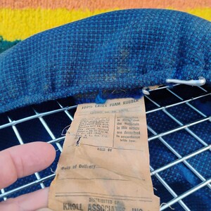 May include: A blue textured fabric cushion attached to a silver metal wire grid frame. A tan paper tag is attached, reading 100% LATEX FOAM RUBBER, DISTRIBUTED BY KNOLL ASSOCIATES, INC, 320 Park Ave., New York, along with legal compliance text.