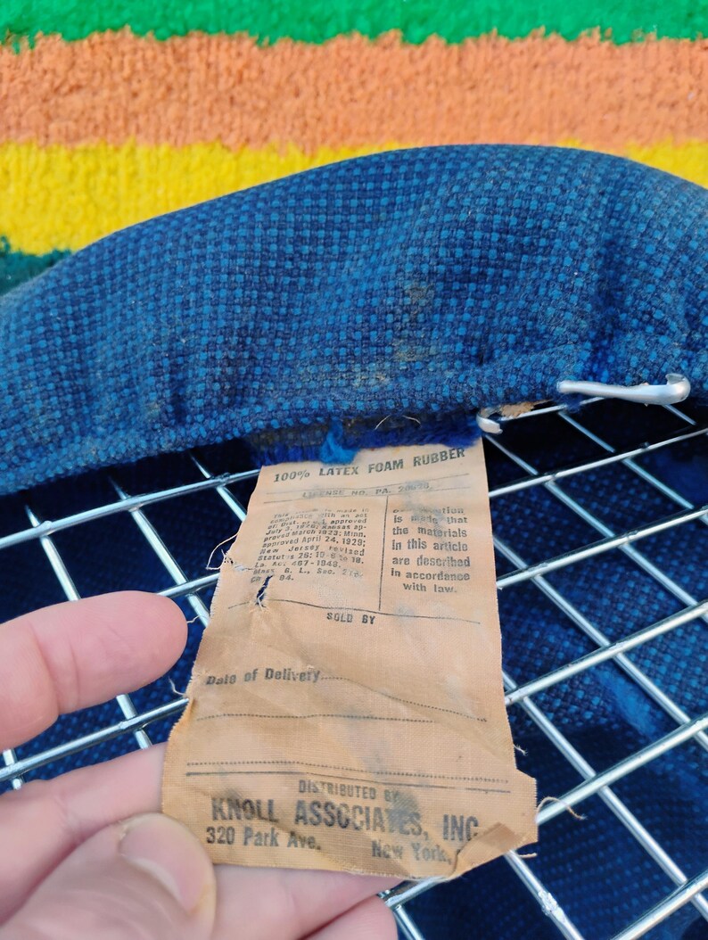 May include: A blue textured fabric cushion attached to a silver metal wire grid frame. A tan paper tag is attached, reading 100% LATEX FOAM RUBBER, DISTRIBUTED BY KNOLL ASSOCIATES, INC, 320 Park Ave., New York, along with legal compliance text.