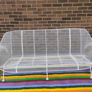 May include: A white metal wire mesh sofa featuring a curved backrest and integrated armrests. The structure is composed of a grid pattern of thin white metal rods, supported by four slender white metal legs, creating a lightweight appearance.