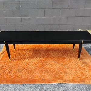 May include: A rectangular black coffee table with a smooth, flat top surface. The table features four tapered black legs, each accented with a metallic gold-toned cap at the base and a small gold-toned detail where the leg meets the table frame.