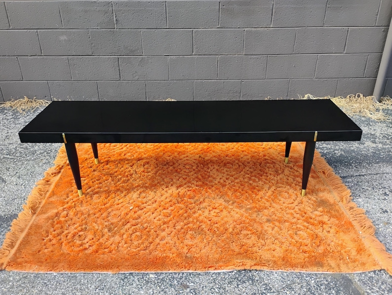 May include: A rectangular black coffee table with a smooth, flat top surface. The table features four tapered black legs, each accented with a metallic gold-toned cap at the base and a small gold-toned detail where the leg meets the table frame.