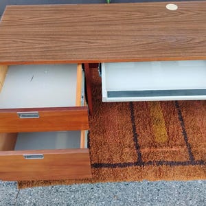 May include: A wooden desk with a brown wood grain top, featuring two stacked drawers on the left side with silver metal handles and a white sliding keyboard tray on the right side. The desk has a circular cutout on the top surface for cable management.