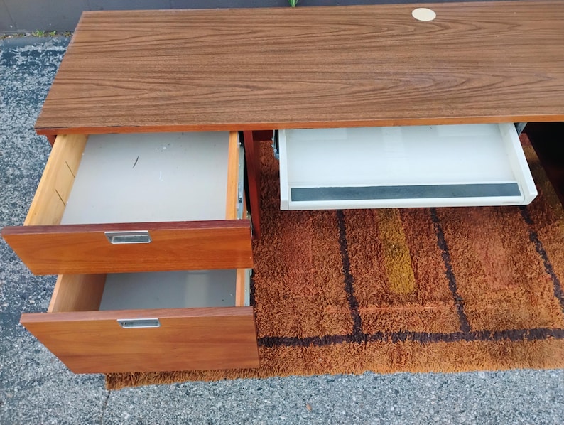 May include: A wooden desk with a brown wood grain top, featuring two stacked drawers on the left side with silver metal handles and a white sliding keyboard tray on the right side. The desk has a circular cutout on the top surface for cable management.