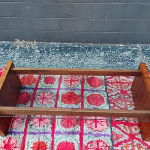 May include: A rectangular wooden coffee table with a clear glass shelf. The table has a warm brown finish and is set on a colorful patterned rug with red, purple, and blue hues. The background is a dark gray wall.