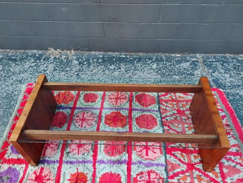 May include: A rectangular wooden coffee table with a clear glass shelf. The table has a warm brown finish and is set on a colorful patterned rug with red, purple, and blue hues. The background is a dark gray wall.