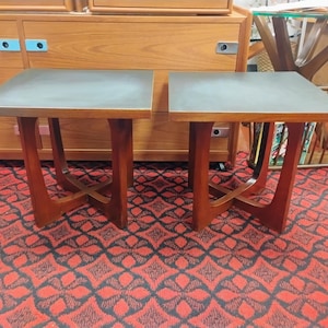 May include: A pair of square side tables featuring dark gray matte tops and warm-toned wooden bases. Each table is supported by four legs that curve inward to meet at a central cross-brace, providing a stable structure for these wooden furniture pieces.