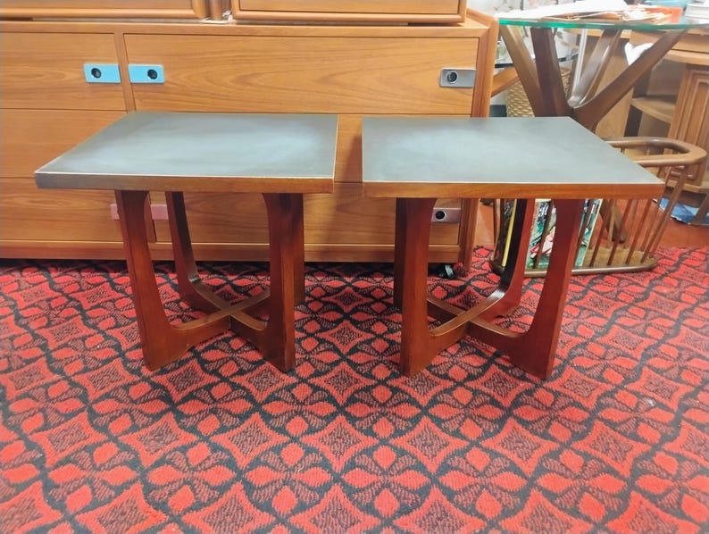 May include: A pair of square side tables featuring dark gray matte tops and warm-toned wooden bases. Each table is supported by four legs that curve inward to meet at a central cross-brace, providing a stable structure for these wooden furniture pieces.