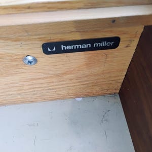 May include: A light-colored wooden furniture component featuring a black rectangular label with the white text "herman miller" and a small logo. A single silver-toned metal screw is visible on the left side of the wood grain surface.