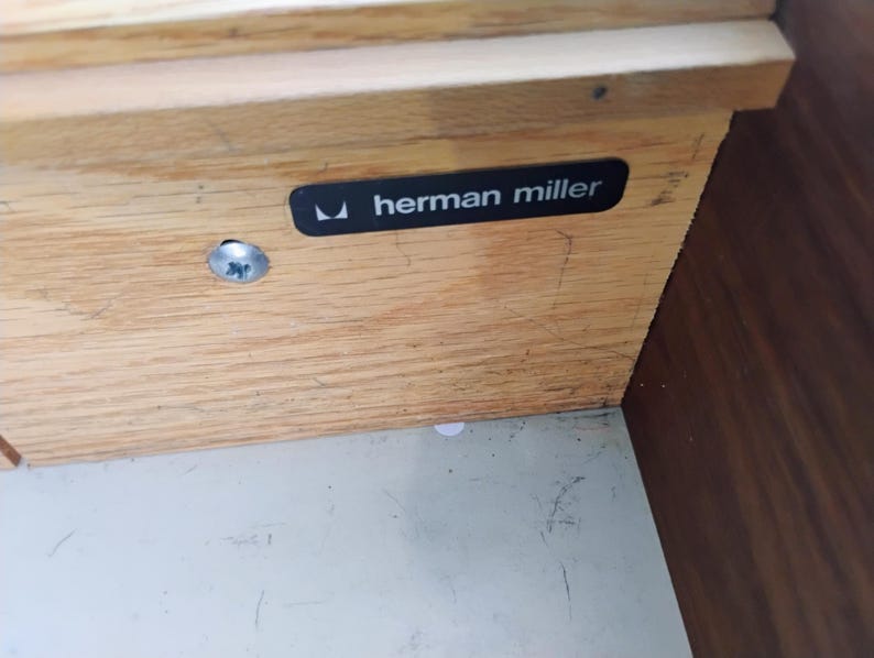 May include: A light-colored wooden furniture component featuring a black rectangular label with the white text "herman miller" and a small logo. A single silver-toned metal screw is visible on the left side of the wood grain surface.