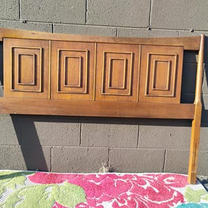 May include: A wooden headboard with a curved top and four rectangular panels with raised edges. The headboard is a light brown color and has a simple, mid-century modern design.