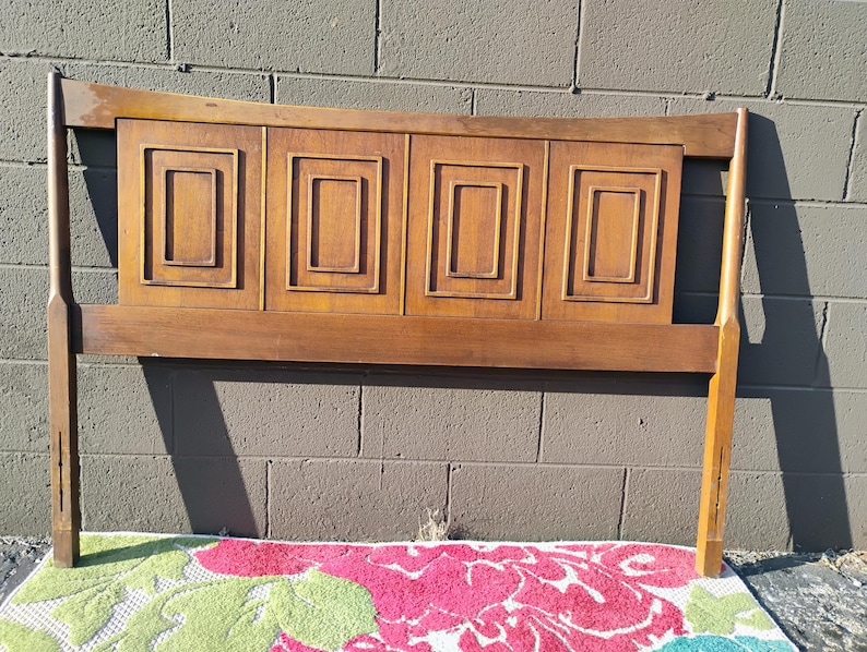 May include: A wooden headboard with a curved top and four rectangular panels with raised edges. The headboard is a light brown color and has a simple, mid-century modern design.