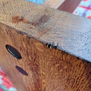 May include: Close-up of a section of a wooden table leg. The wood grain is visible, with a warm brown color and a few dark oval-shaped holes. The edges show some wear and tear.