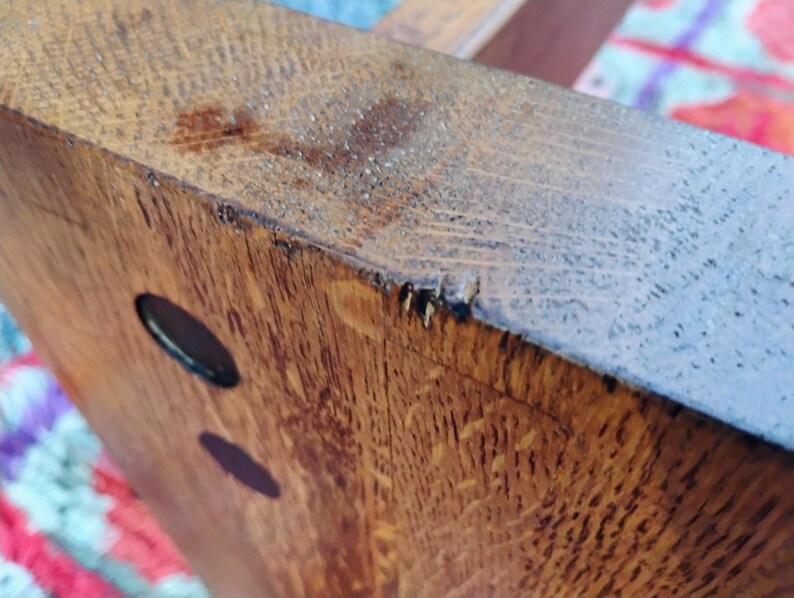 May include: Close-up of a section of a wooden table leg. The wood grain is visible, with a warm brown color and a few dark oval-shaped holes. The edges show some wear and tear.