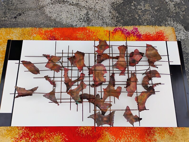 May include: A metal wall sculpture with a grid pattern and abstract shapes. The shapes are made of copper and have a reddish-brown color.