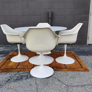 May include: A white dining set consisting of a round table and four matching chairs. Each chair features a curved shell seat supported by a single central pedestal base. The table also stands on a matching white central pedestal base.
