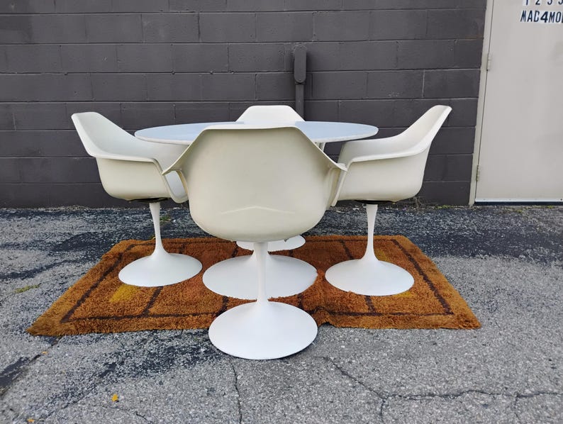 May include: A white dining set consisting of a round table and four matching chairs. Each chair features a curved shell seat supported by a single central pedestal base. The table also stands on a matching white central pedestal base.