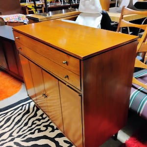 Puede incluir: Un armario de madera vintage con dos cajones y cuatro puertas. El armario tiene un acabado en madera clara y herrajes de lat&oacute;n. El armario est&aacute; sobre una alfombra de estampado de cebra.