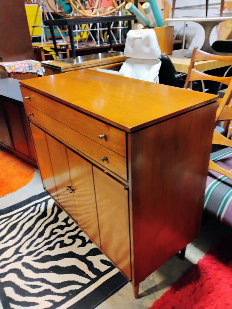 Puede incluir: Un armario de madera vintage con dos cajones y cuatro puertas. El armario tiene un acabado en madera clara y herrajes de lat&oacute;n. El armario est&aacute; sobre una alfombra de estampado de cebra.