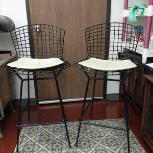 May include: Two black wire frame bar stools with white padded seats. The stools are on a patterned rug in a room with a wooden door and a shelf unit in the background.