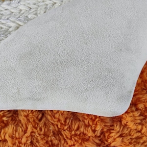May include: Close-up of a textured, off-white fabric with a curved edge, resting on a vibrant orange, shaggy material. A section of white and silver knitted fabric is visible in the upper left corner.