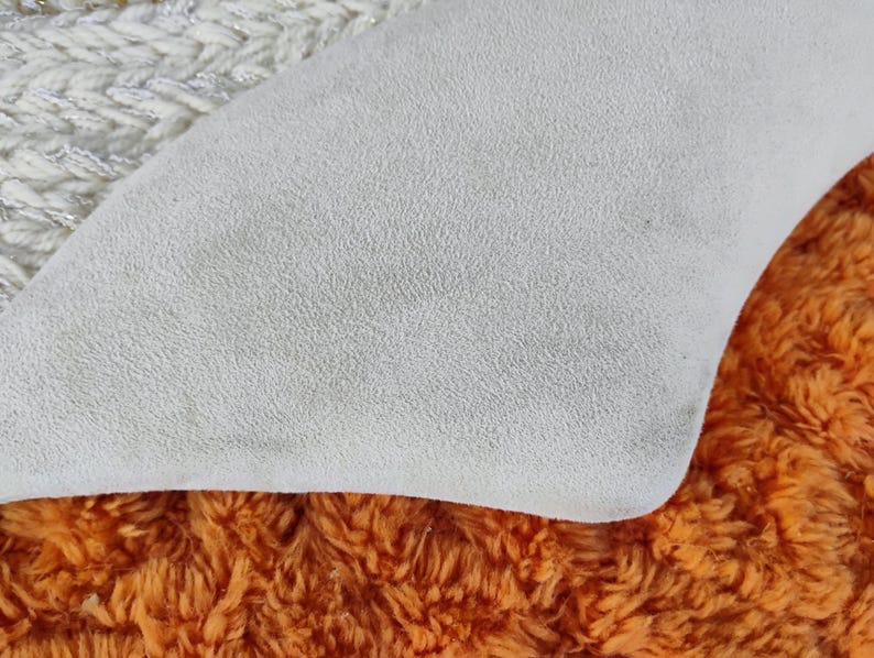 May include: Close-up of a textured, off-white fabric with a curved edge, resting on a vibrant orange, shaggy material. A section of white and silver knitted fabric is visible in the upper left corner.