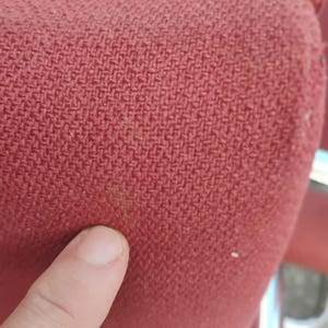 May include: A close-up view of a textured, woven fabric in a deep red color. The material features a distinct cross-hatch pattern with a visible weave structure. A small, faint brownish discoloration is present on the surface of the red textile.