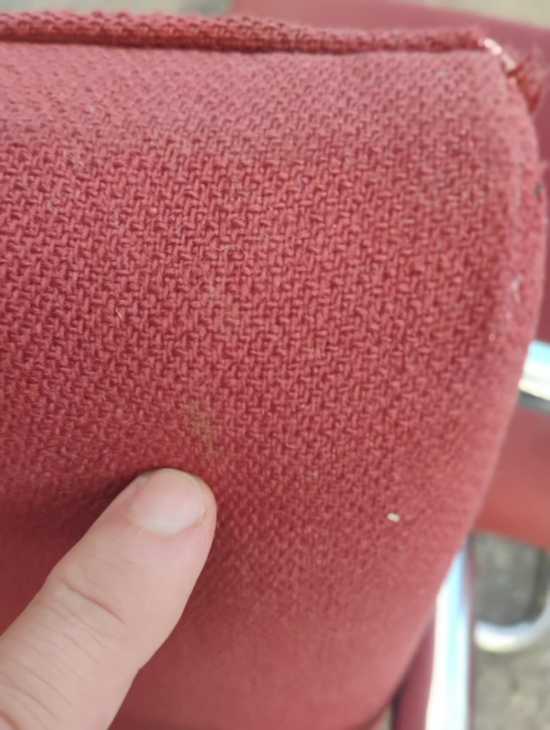May include: A close-up view of a textured, woven fabric in a deep red color. The material features a distinct cross-hatch pattern with a visible weave structure. A small, faint brownish discoloration is present on the surface of the red textile.