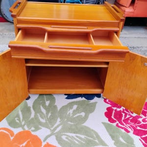 May include: A wooden serving cart with two doors, a drawer, and a top shelf. The cart has wheels and is made of light wood.