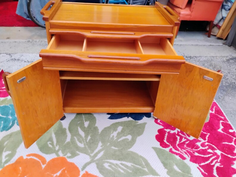 May include: A wooden serving cart with two doors, a drawer, and a top shelf. The cart has wheels and is made of light wood.