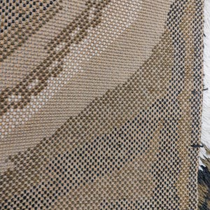May include: Close-up of a woven fabric with a diagonal pattern in shades of brown, black, and white. The fabric has a textured surface and a slightly frayed edge.