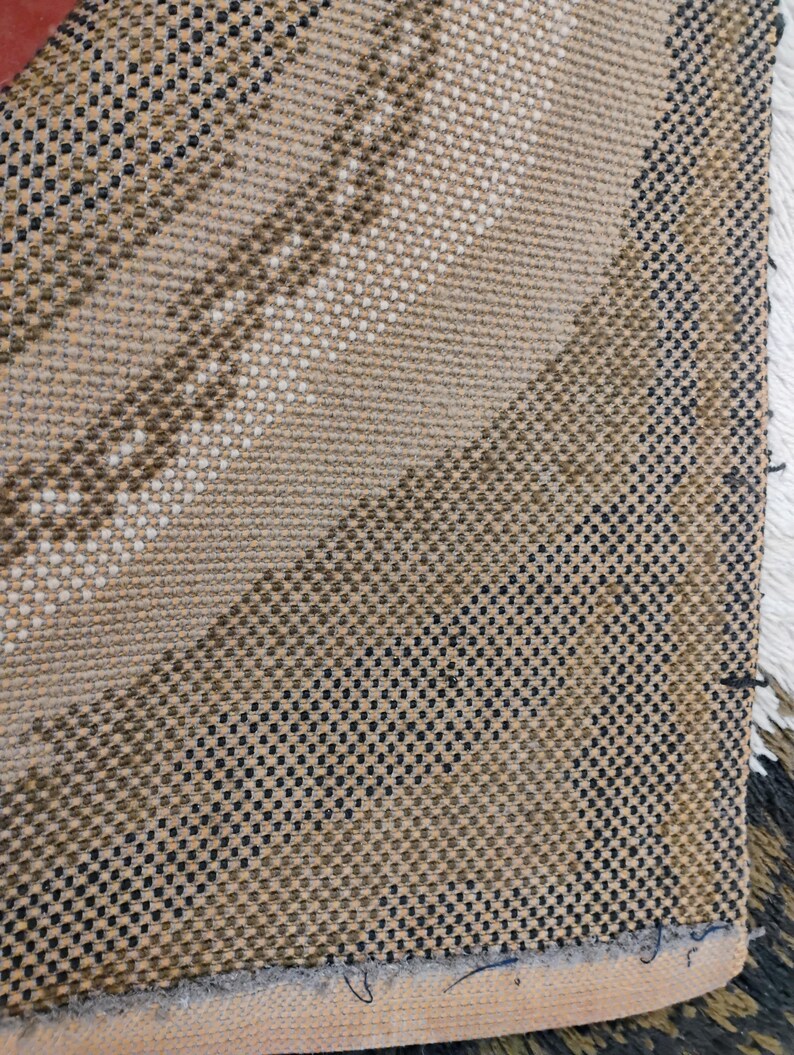 May include: Close-up of a woven fabric with a diagonal pattern in shades of brown, black, and white. The fabric has a textured surface and a slightly frayed edge.