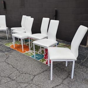 May include: A set of six white dining chairs with a minimalist design. The chairs have white seats and backs, supported by white metal frames. They are arranged on a floral patterned rug, set against a dark gray brick wall.
