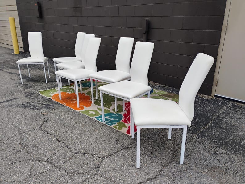 May include: A set of six white dining chairs with a minimalist design. The chairs have white seats and backs, supported by white metal frames. They are arranged on a floral patterned rug, set against a dark gray brick wall.