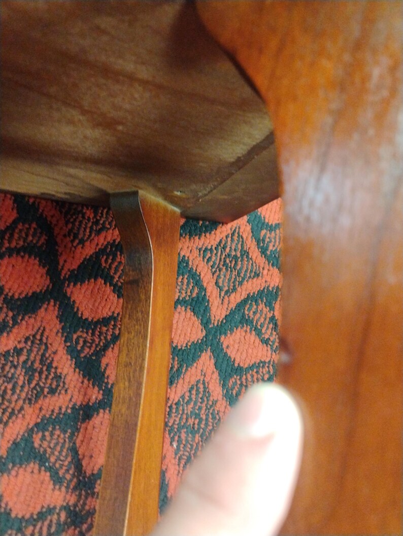 May include: A wooden furniture leg with a curved design attached to a flat wooden surface. The wood has a medium brown tone with a visible grain pattern. The leg connects to the underside of the furniture piece at a right angle.
