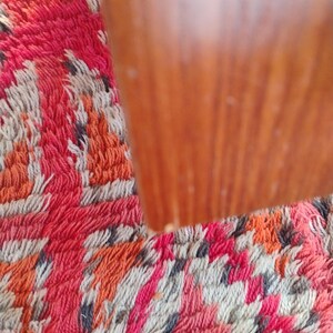 May include: Close-up of a wooden object with a warm, reddish-brown hue. The wood grain is visible. The object is set against a colorful rug with a pattern of red, orange, white, and gray.