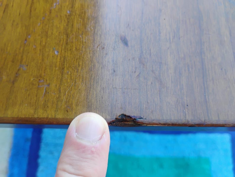 May include: Close-up of a wooden table top with a small chip or crack in the finish. The table top is a light brown color and the chip is a darker brown.
