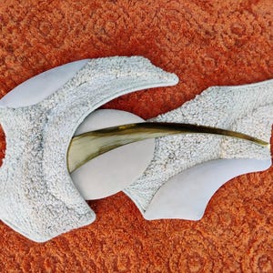 May include: Abstract sculpture featuring a light grey, textured, fabric-like material and smooth, white, curved elements. A curved, horn-like piece is centered. The sculpture is set against an orange background.