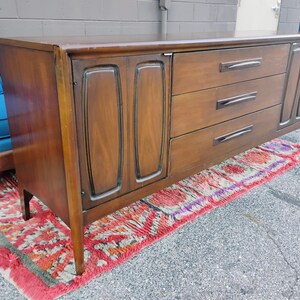 May include: A long, rectangular wooden sideboard featuring a dark brown finish. The piece includes a cabinet section with two doors on the left, three horizontal drawers with dark handles in the center, and a single cabinet door on the right side.