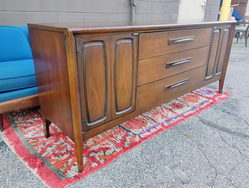 May include: A long, rectangular wooden sideboard featuring a dark brown finish. The piece includes a cabinet section with two doors on the left, three horizontal drawers with dark handles in the center, and a single cabinet door on the right side.