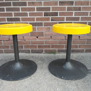 May include: Two yellow round stools with black bases. The stools are made of plastic and have a simple, modern design.