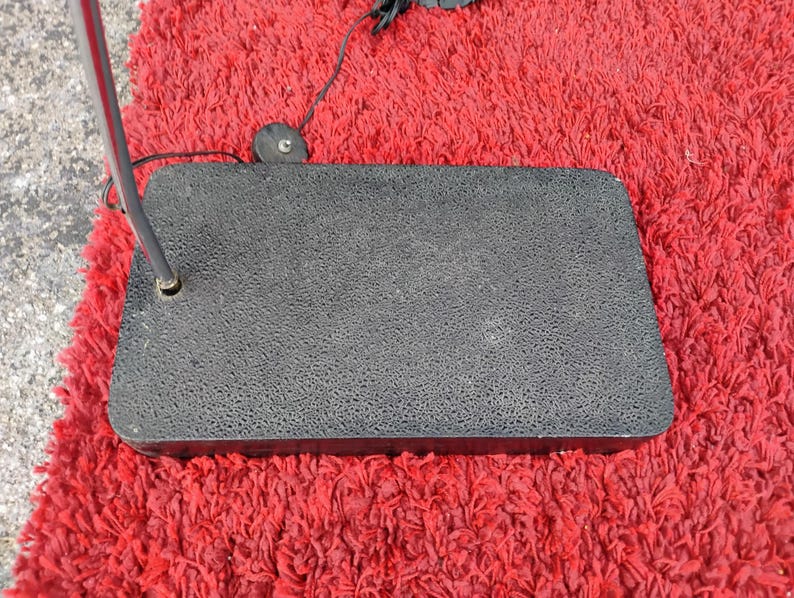 K&ouml;nnte beinhalten: Ein schwarzer, rechteckiger Sockel mit strukturierter Oberfl&auml;che, m&ouml;glicherweise f&uuml;r eine Lampe oder ein &auml;hnliches Ger&auml;t. Eine Metallstange erstreckt sich von der Basis, und ein roter, zotteliger Teppich bildet den Hintergrund.