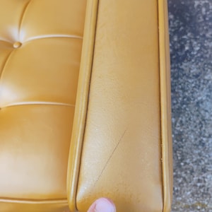 May include: A mustard yellow upholstered sofa armrest featuring a tufted seat cushion with visible buttons. The armrest surface displays a thin, dark linear scratch mark running diagonally across the smooth, leather-like material texture.