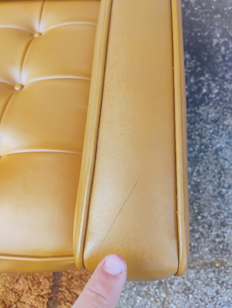 May include: A mustard yellow upholstered sofa armrest featuring a tufted seat cushion with visible buttons. The armrest surface displays a thin, dark linear scratch mark running diagonally across the smooth, leather-like material texture.