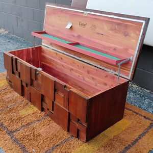 Puede incluir: Un arc&oacute;n de cedro vintage con tapa abatible y bandeja incorporada. El arc&oacute;n est&aacute; hecho de madera de color marr&oacute;n rojizo y tiene un dise&ntilde;o geom&eacute;trico en la parte delantera. El interior est&aacute; forrado con cedro. El arc&oacute;n est&aacute; abierto, revelando el interior.