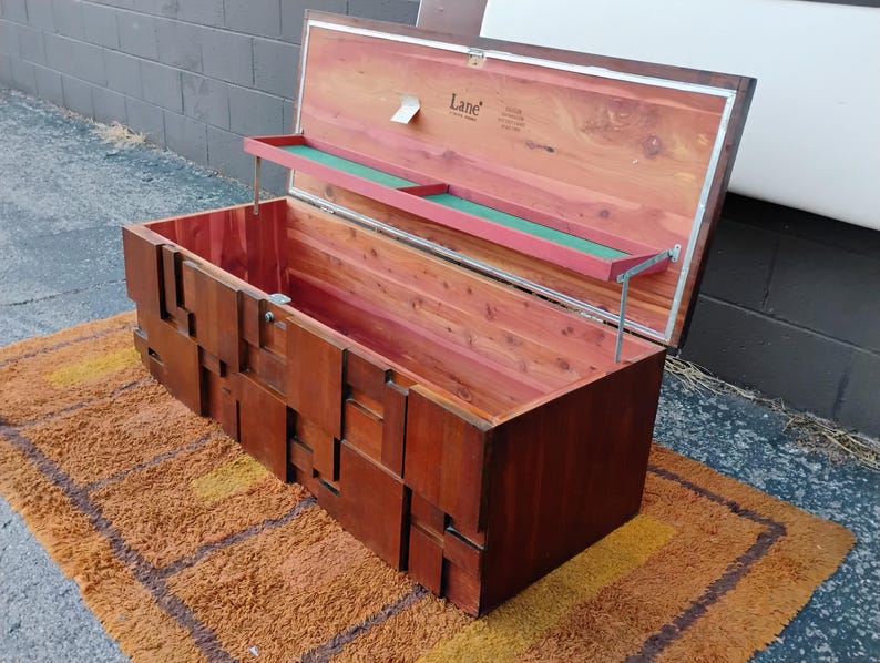 Puede incluir: Un arc&oacute;n de cedro vintage con tapa abatible y bandeja incorporada. El arc&oacute;n est&aacute; hecho de madera de color marr&oacute;n rojizo y tiene un dise&ntilde;o geom&eacute;trico en la parte delantera. El interior est&aacute; forrado con cedro. El arc&oacute;n est&aacute; abierto, revelando el interior.
